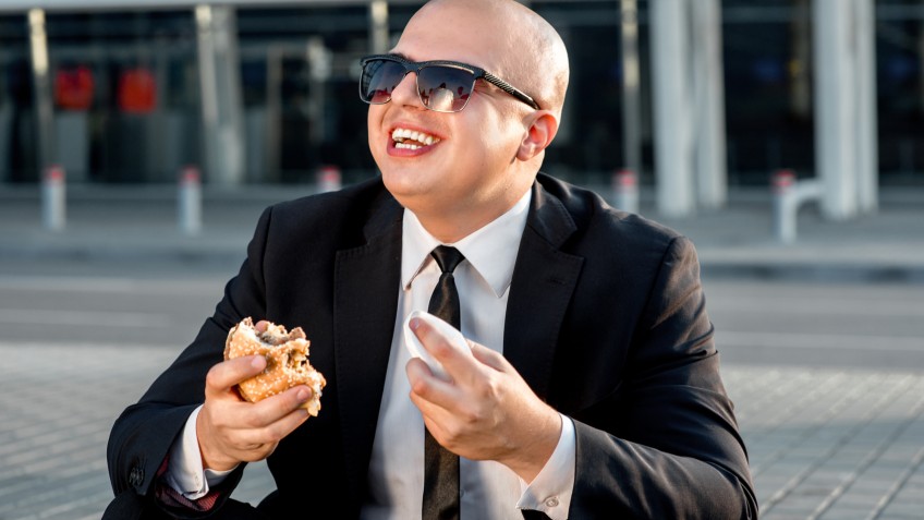 Businessman or banker working with laptop and having snack with burger outside the airport or contemporary building