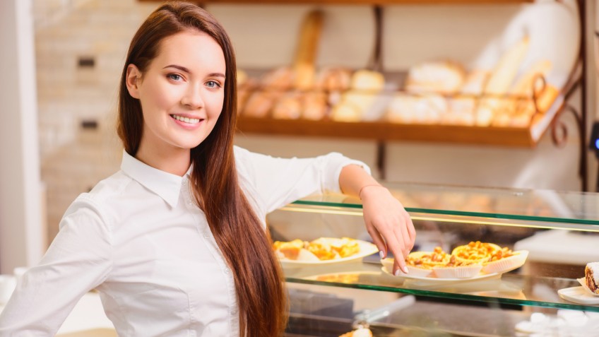 Proud of what we have created. Beautiful young female baker standing by the windowsill of her bakery and pointing at a variety of desserts and rows of fresh baked bread