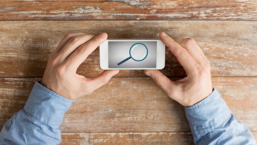 business, education, people and technology concept - close up of male hands holding smartphone with magnifying glass picture on screen at table