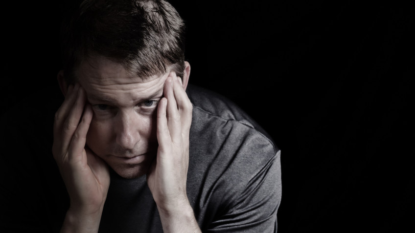 Closeup front view of mature man with head down, touching his temples, while displaying depression on black background