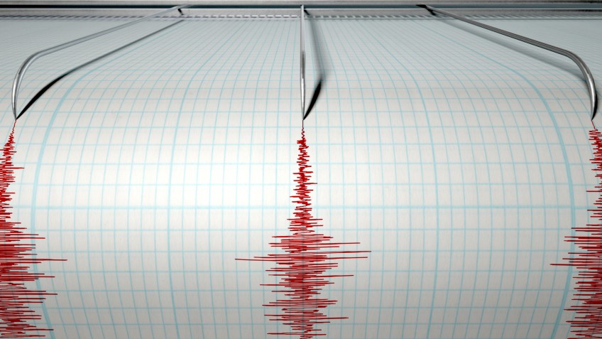 A closeup of a seismograph machine needle drawing a red line on graph paper depicting seismic and eartquake activity on an isolated white background