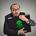 Money greed. Business man holding holding case with dollars tightly isolated on grey wall background. Worship, miser, excessive gain, finance concept
